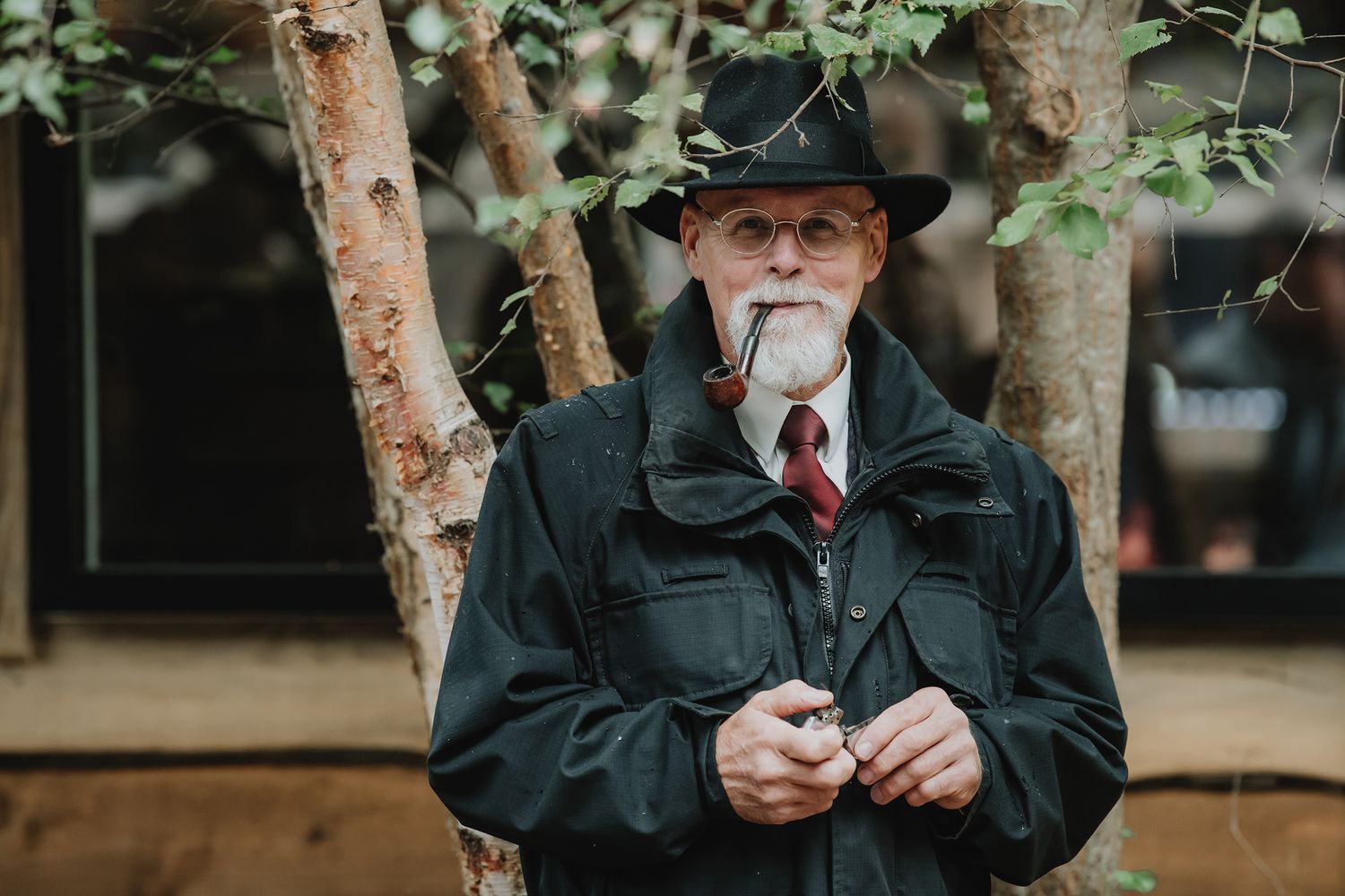 An elderly man with a pipe and black hat stands outdoors among trees.