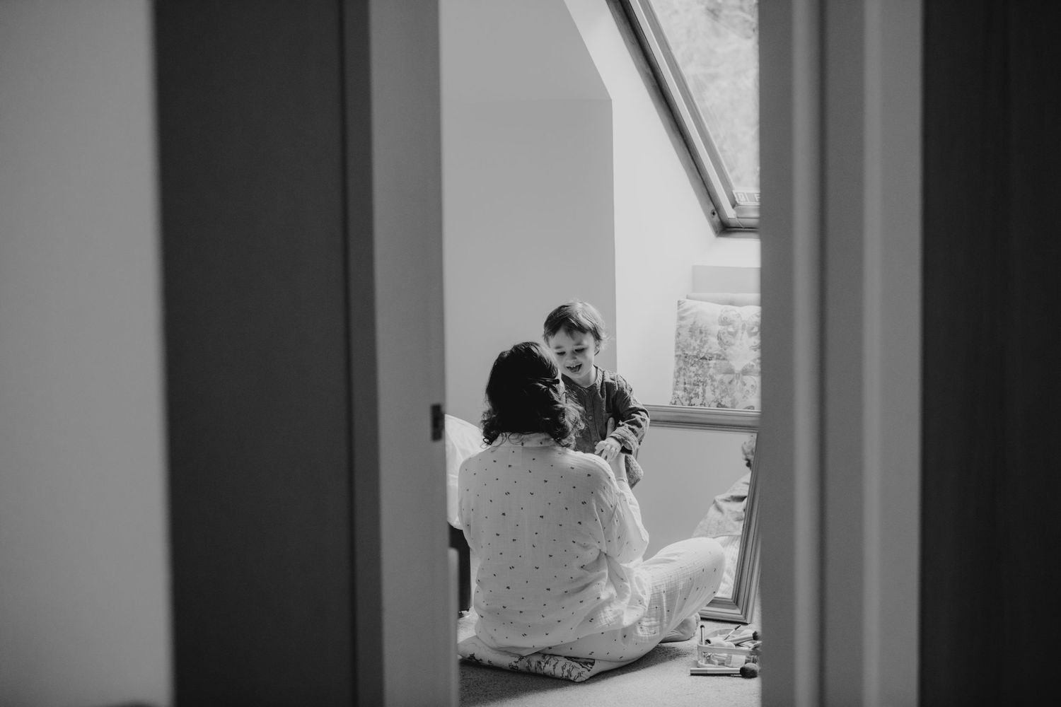 Black and white image of bride sharing a joyful moment with a child in a sunlit attic room.