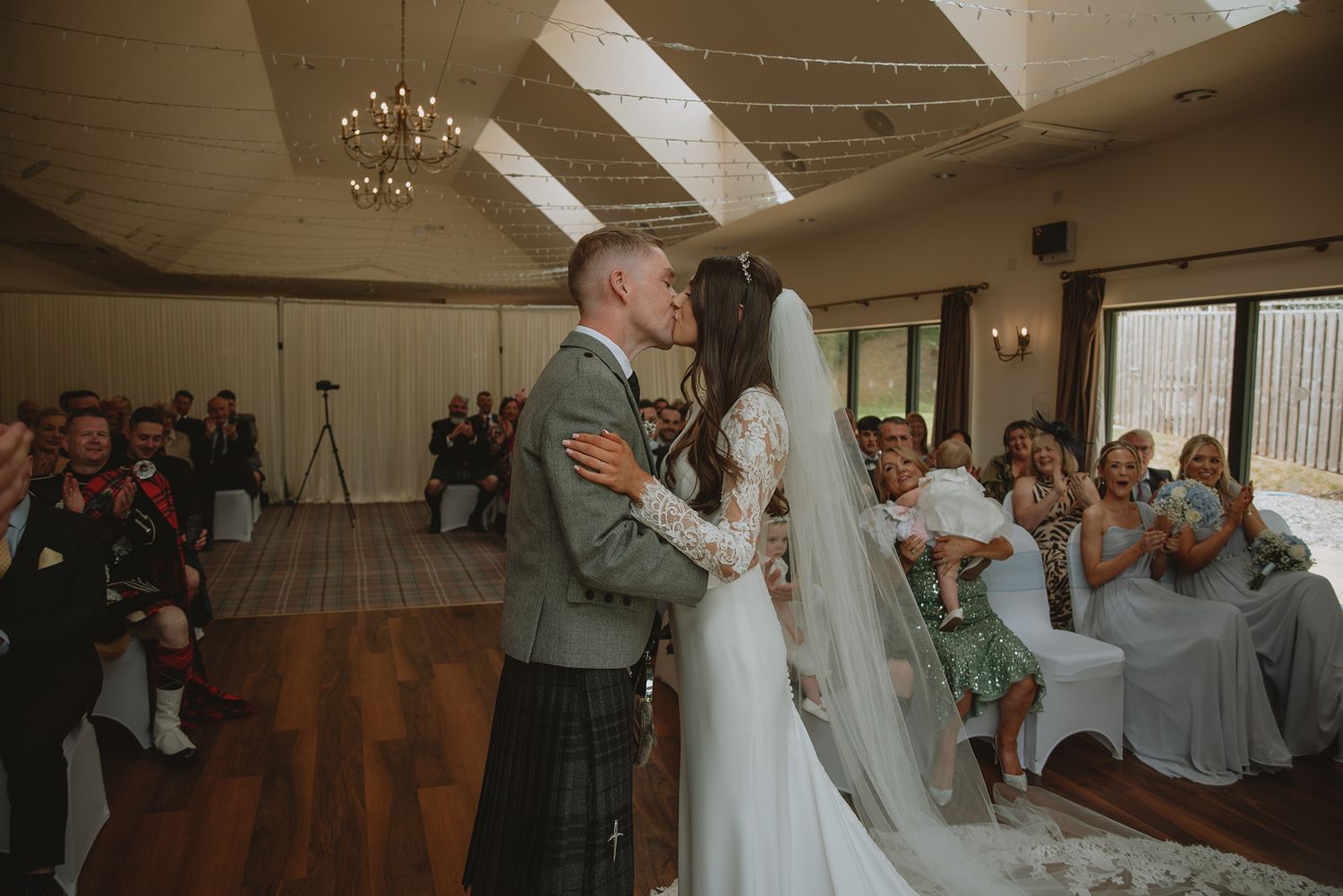 Bride and groom share first kiss at rustic wedding venue with guests seated in background.