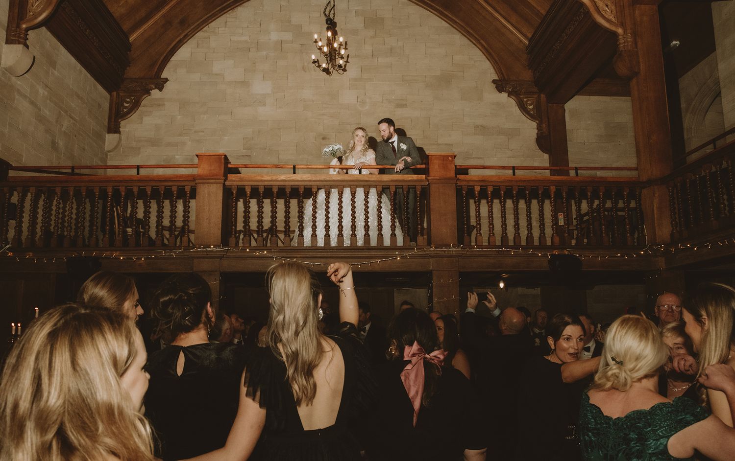 Couple stands on balcony overlooking wedding reception guests dancing in elegant stone venue.