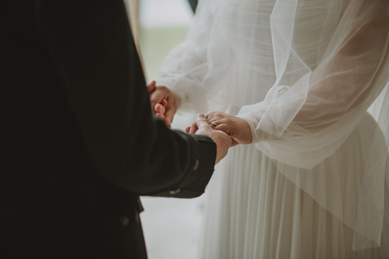 Close-up of bride and groom holding hands during wedding ceremony, bride in elegant white lace gown.