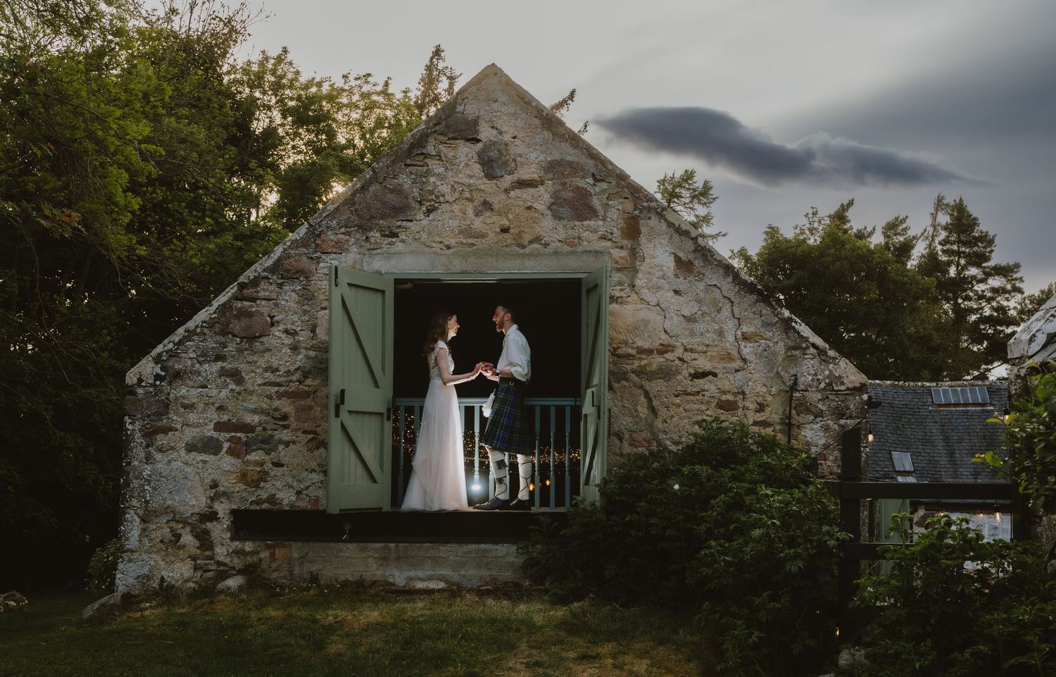 Bride and groom standing in doorway of charming rustic stone building with green shutters at twilight.