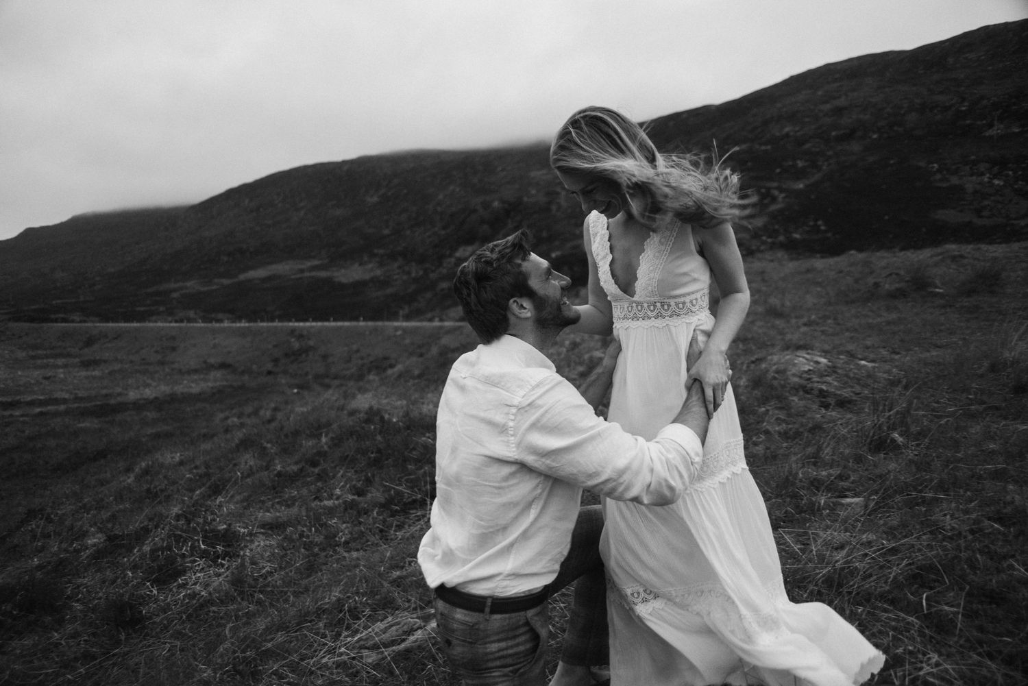 Black and white photo of groom kneeling before bride in flowing dress on misty Scottish hillside.