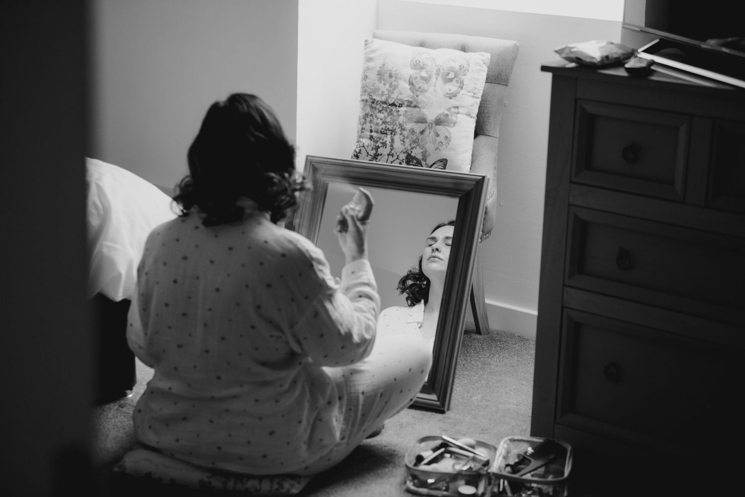 Black and white photo of bride applying makeup while seated before a mirror in a bedroom.