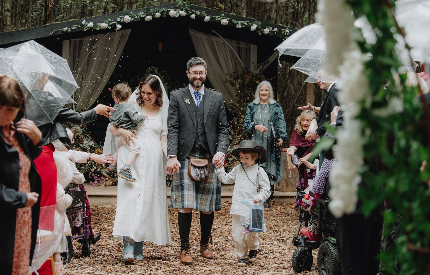 Bride and groom in tartan kilt walk with children through woodland wedding venue as guests hold umbrellas.