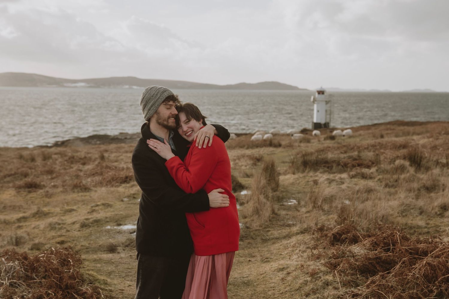 Couple embracing on rugged coastal landscape with gray skies, lighthouse in background, woman in red dress.