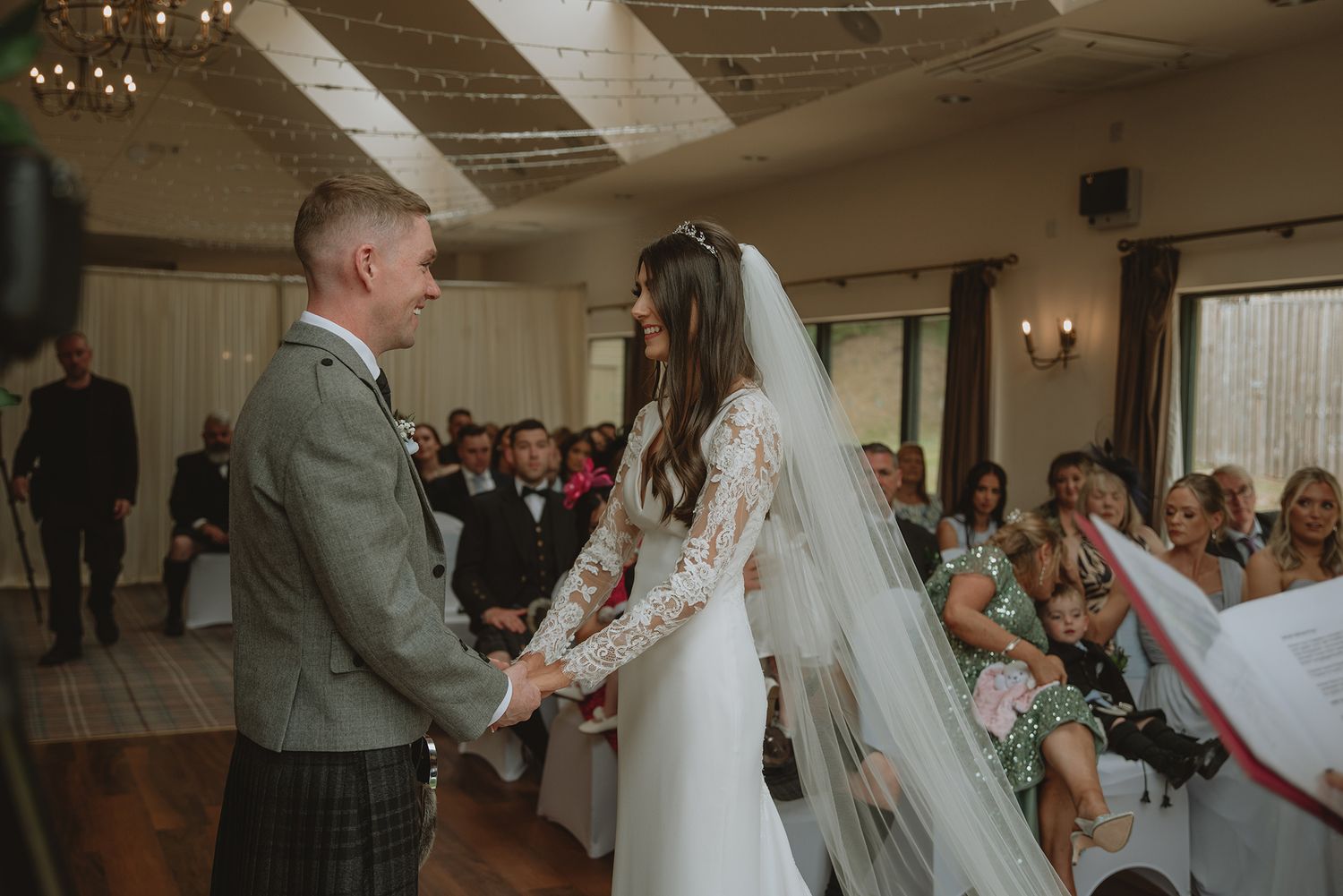 Bride and groom holding hands at wedding ceremony in rustic venue, bride wearing long veil and lace dress, groom in tartan kilt.