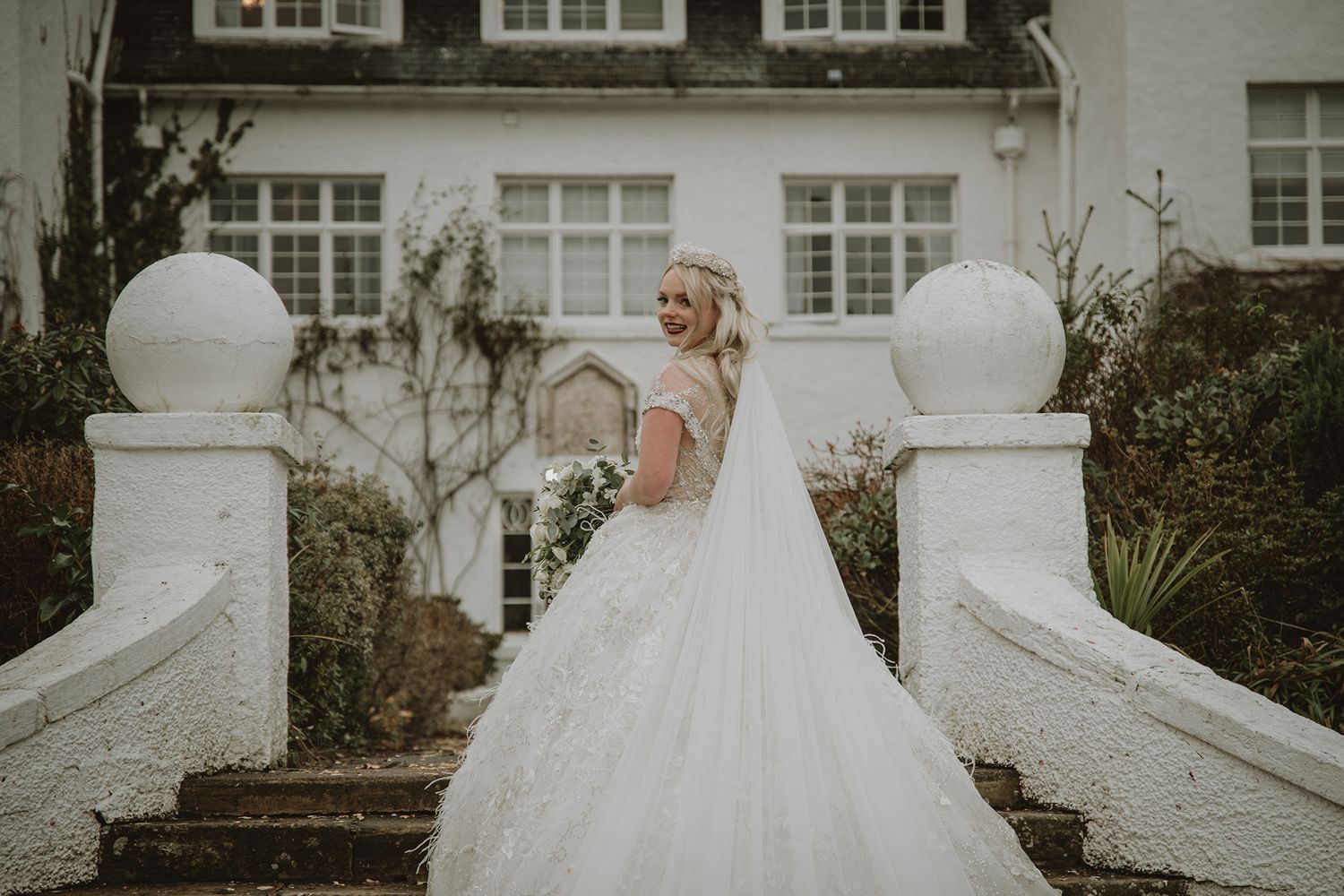 Bride in elegant white ball gown with lace back poses on stone steps before a grand manor house.