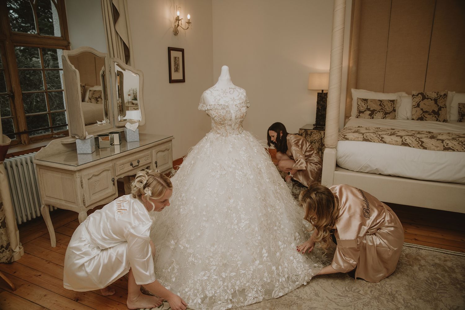 Bridesmaids in satin robes adjusting an ornate white ball gown wedding dress on a mannequin in an elegant room.