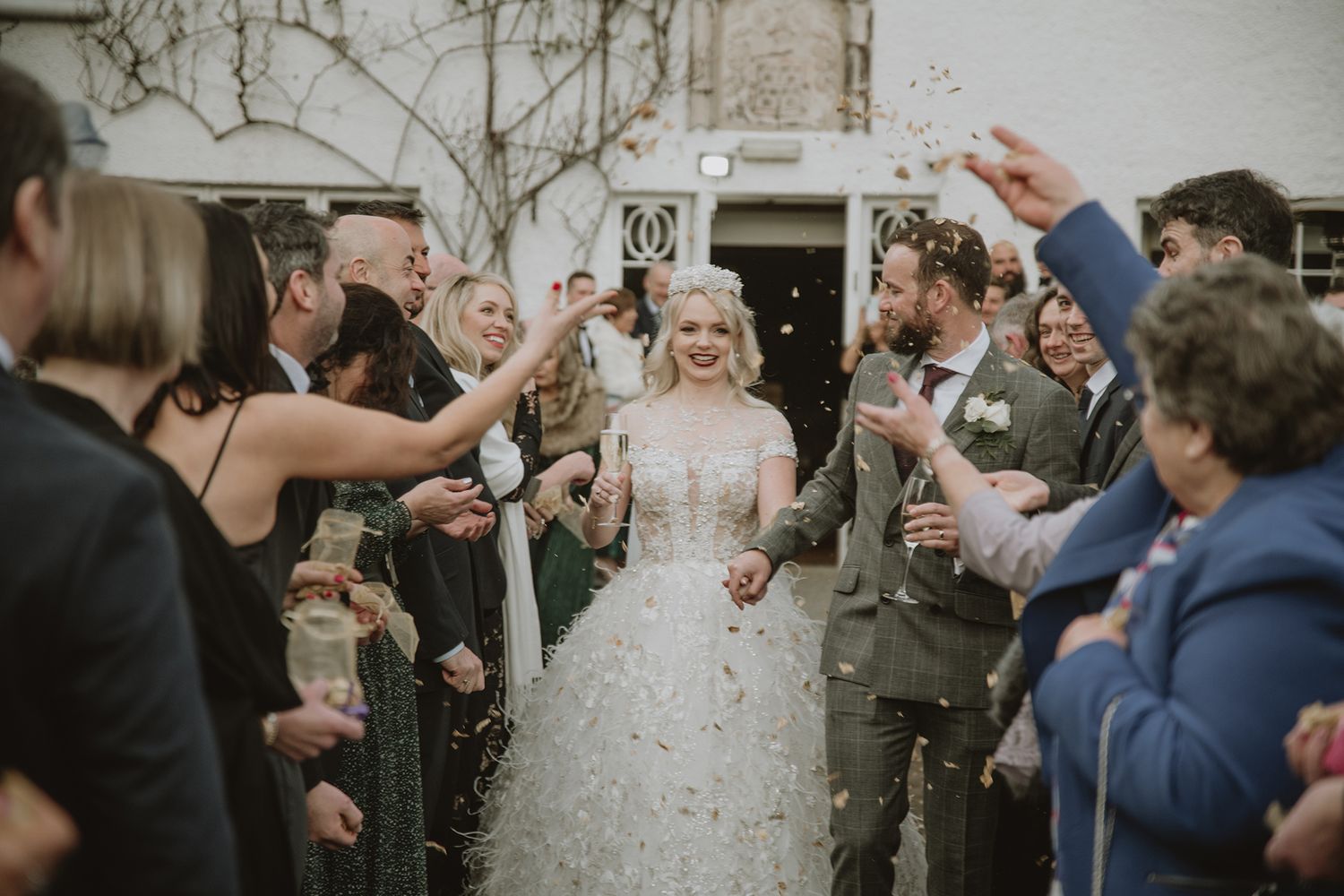 Bride and groom walk through confetti shower surrounded by cheering guests outside a stone venue.