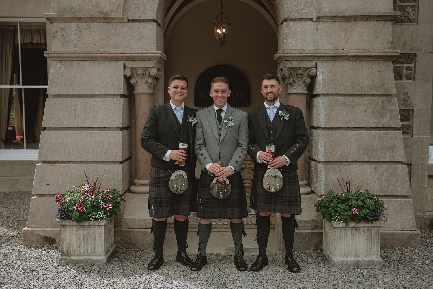 Three men in traditional Scottish kilts and sporrans pose together outside a grand stone building.