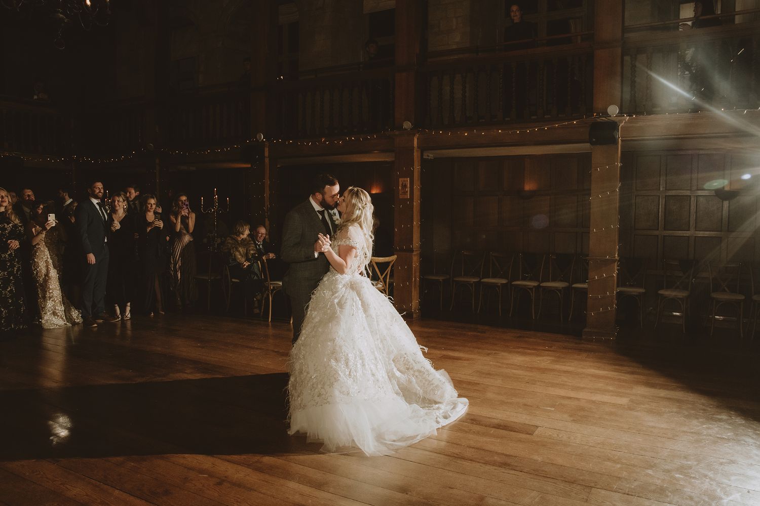 Bride and groom share first dance in elegant wooden ballroom with guests watching in warm romantic lighting.