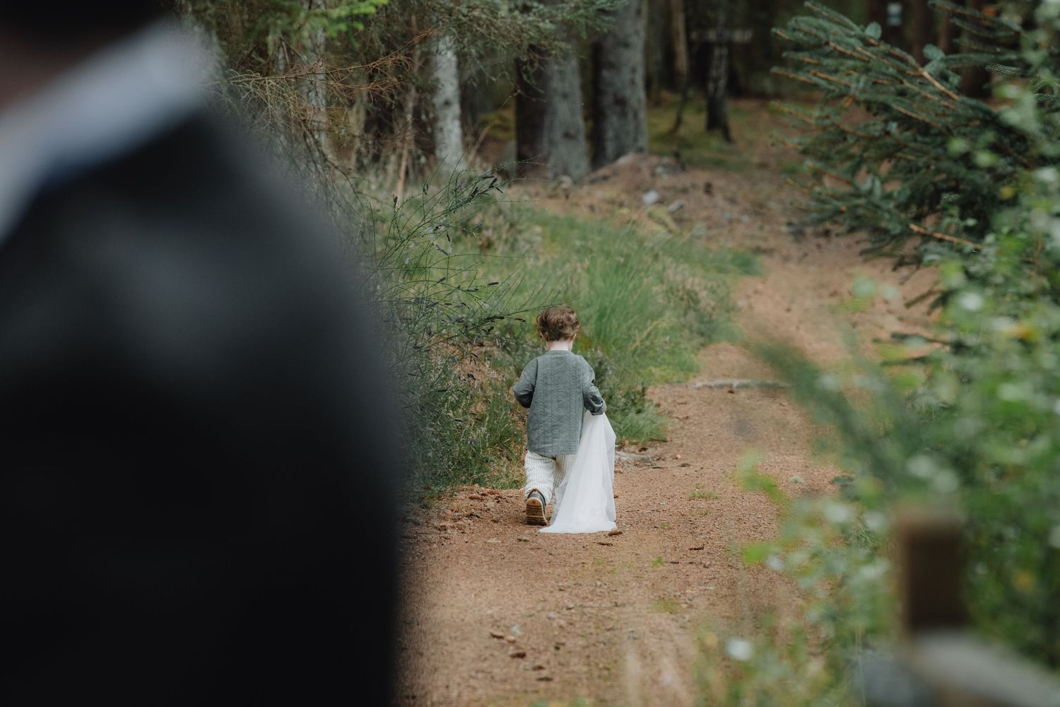 Young child in formal attire runs along woodland dirt path at outdoor forest wedding celebration.