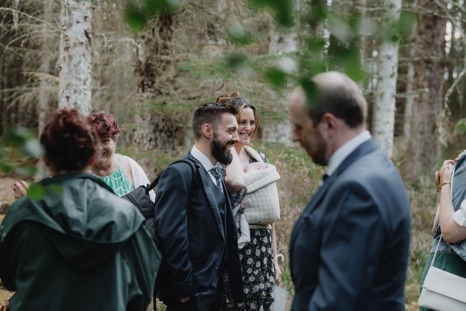 Bearded groom smiles warmly while chatting with guests at relaxed outdoor woodland wedding reception.