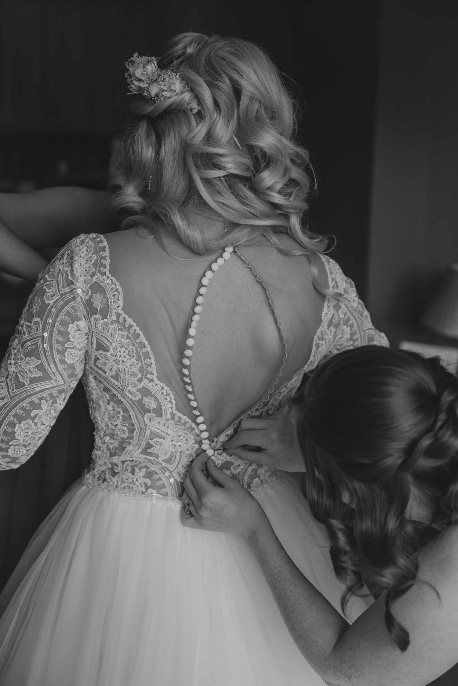 Black and white close-up of intricate lace button-back wedding dress being fastened by a bridesmaid.