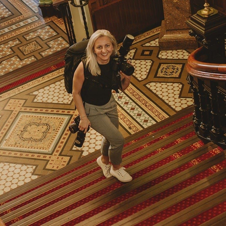 Female photographer with cameras smiling on ornate tiled floor of a grand historic building shot from above.