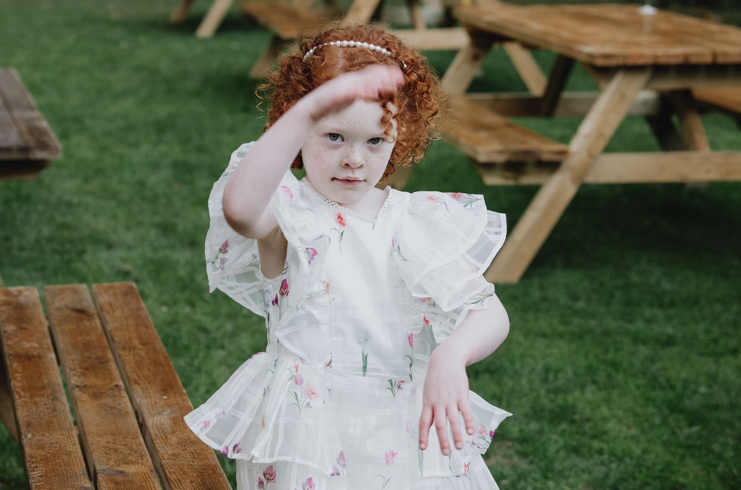 A curly red-haired girl in a floral white dress plays on a green lawn.