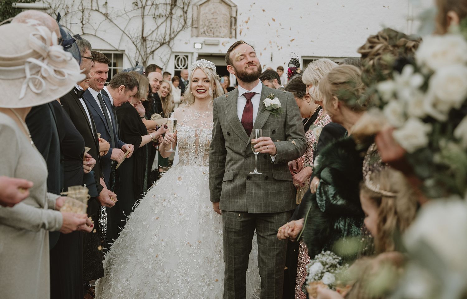 Bride and groom walk through confetti shower surrounded by wedding guests outdoors at a rustic venue celebration.