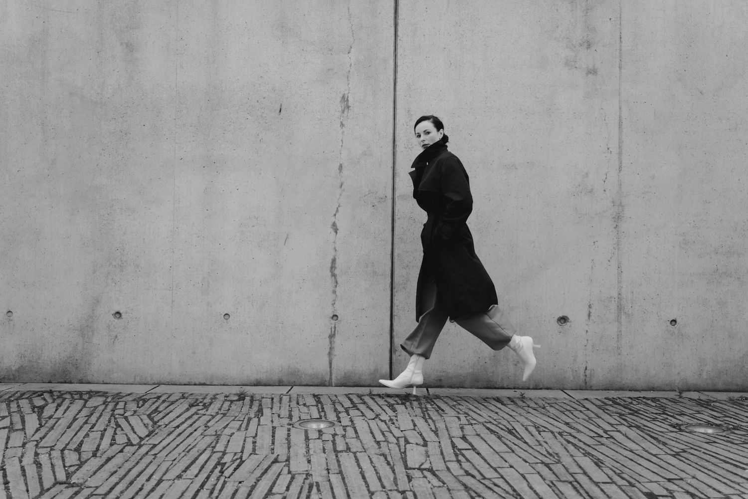 Black and white photo of a woman in a long black coat walking confidently along a cobblestone street beside a concrete wall.