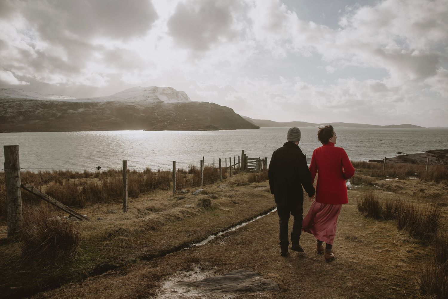 Couple walking hand in hand along coastal path overlooking dramatic Scottish loch scenery.