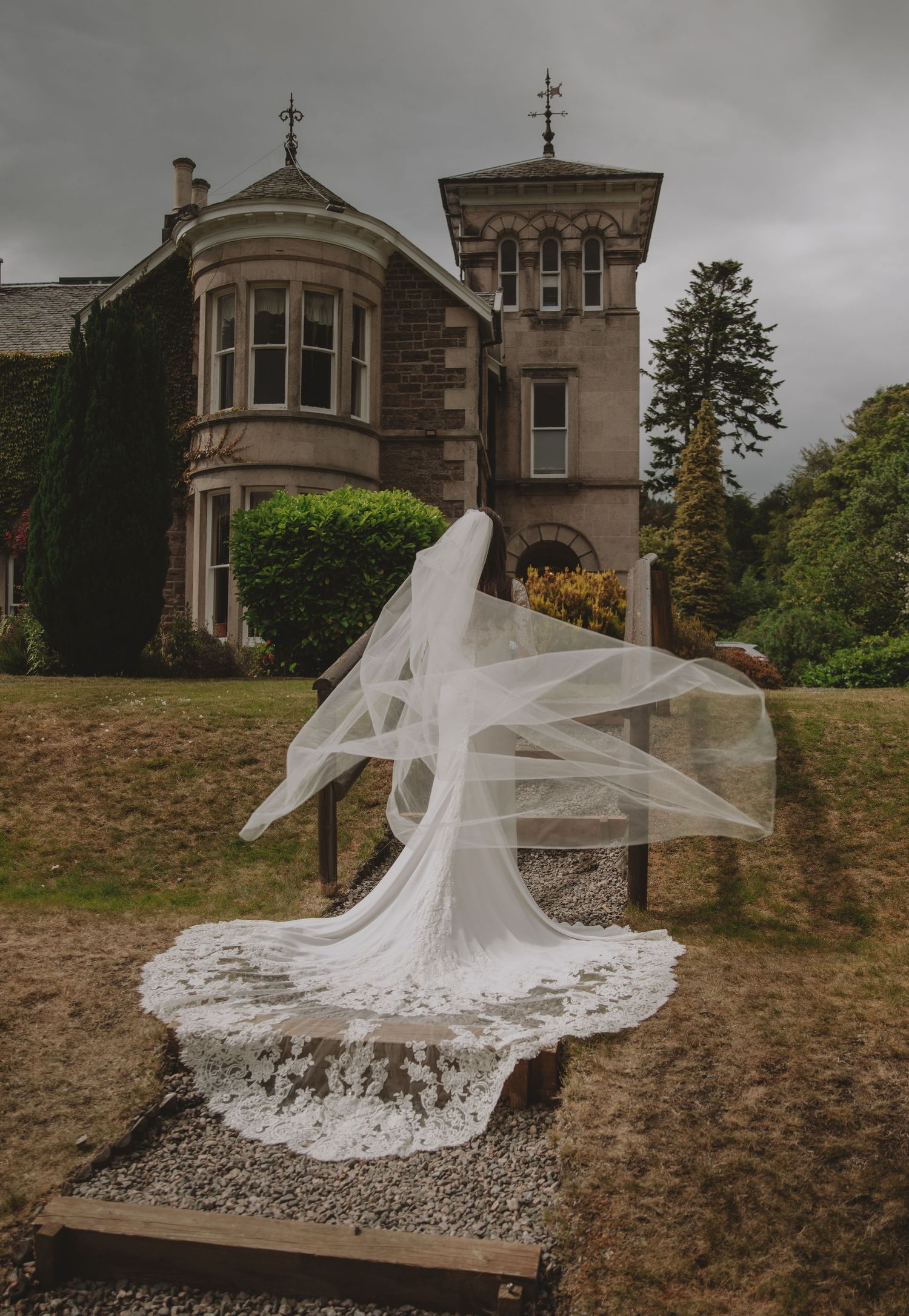 Bride in flowing white gown and dramatic veil billowing in wind before a historic stone manor house on overcast day.