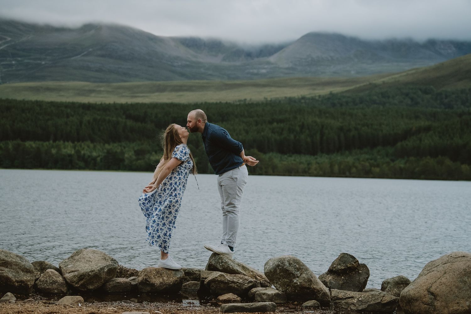 Couple dancing on rocks by a scenic mountain lake with lush green forests in the background.