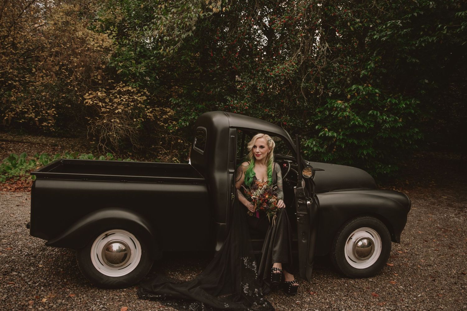 Bride in a dramatic black wedding gown posing beside a vintage matte black pickup truck in an autumn forest.
