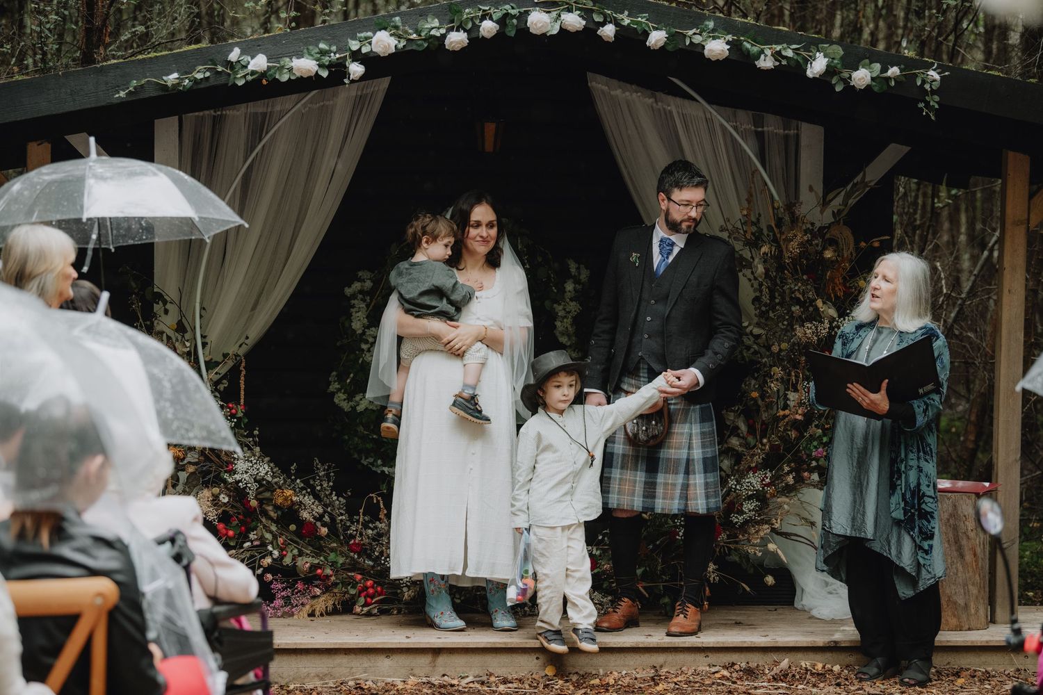 Scottish wedding couple poses with children and officiant outside decorated woodland gazebo after ceremony.