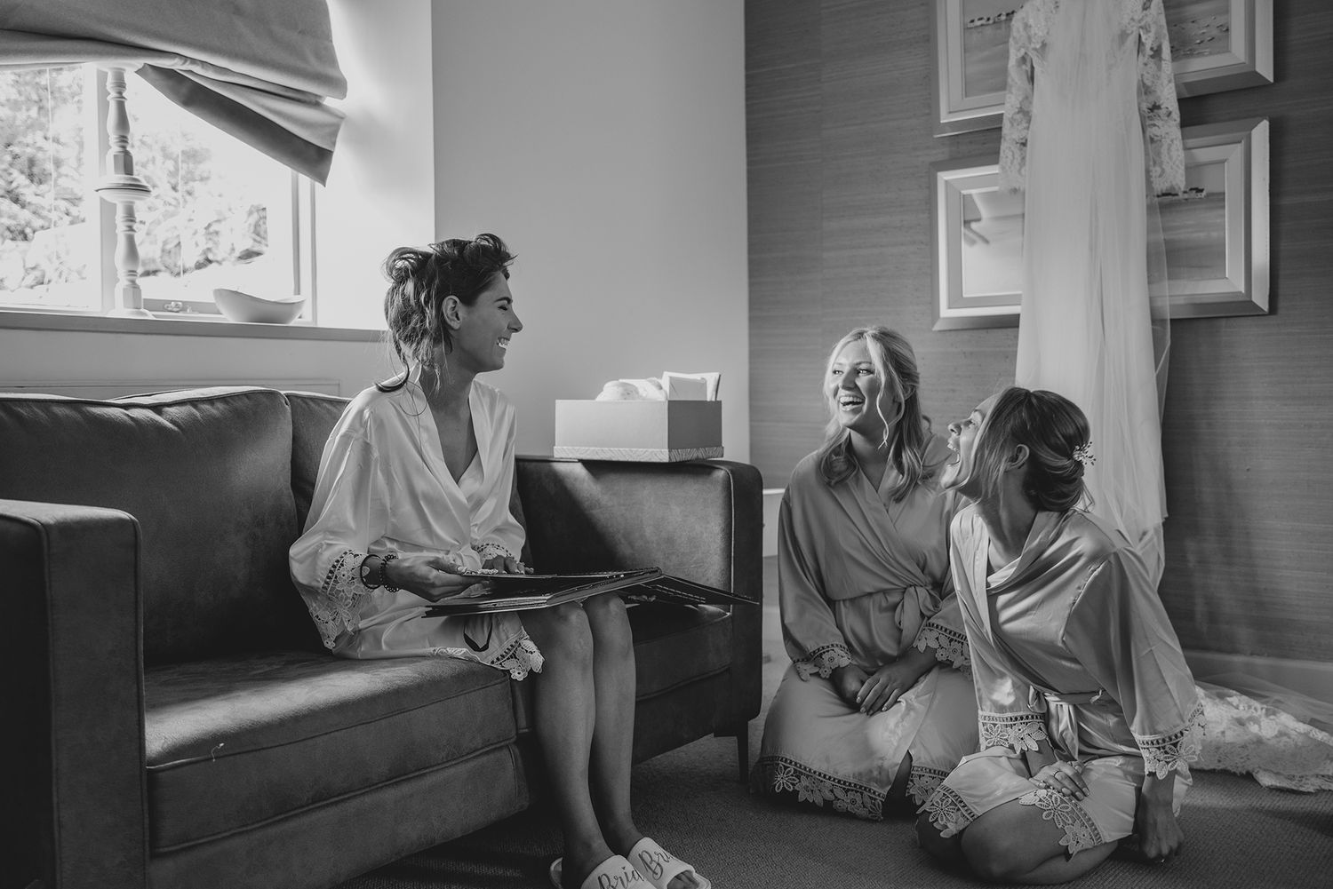 Three bridesmaids in white robes relaxing in a hotel room during wedding preparations, black and white photo.