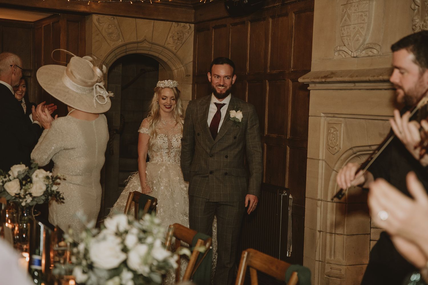 Bride and groom enter wedding reception in elegant wood-paneled hall, guests applauding, floral centerpieces in foreground.
