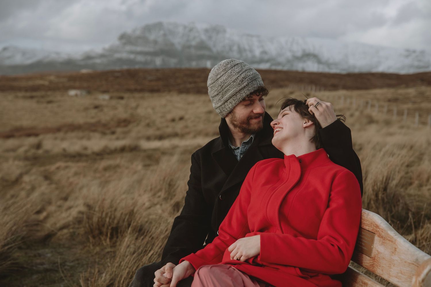 Couple sharing a romantic moment outdoors in a snowy mountain landscape, woman in red hoodie leaning on man in gray beanie.