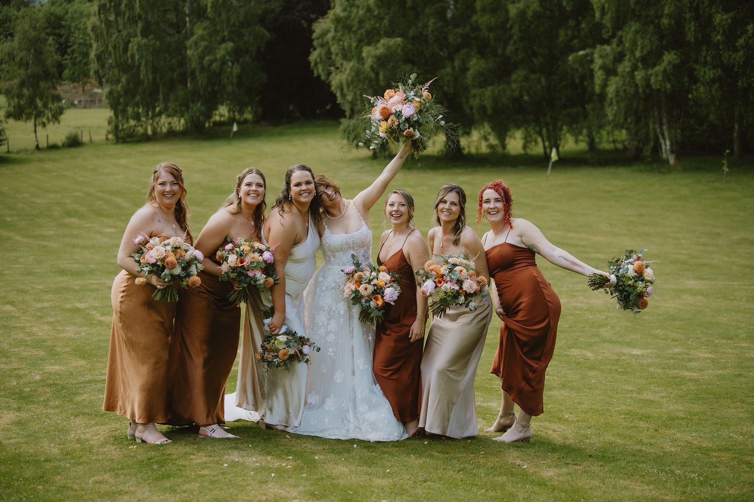 Bride with bridesmaids in earth-tone satin dresses holding bouquets on green lawn.