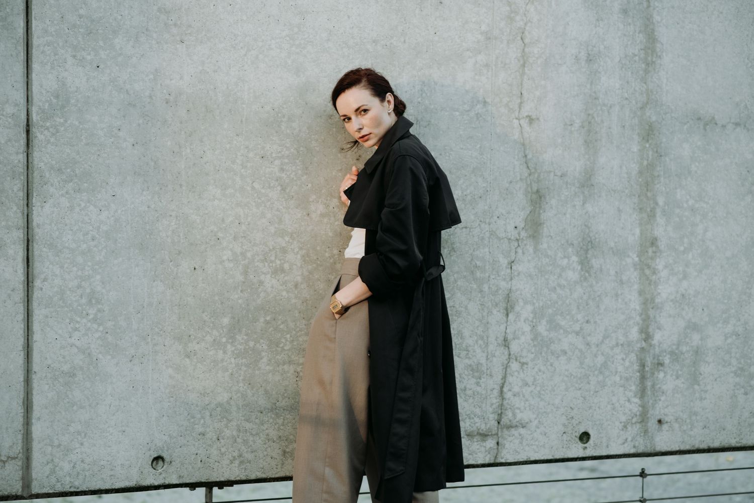 Fashionable woman in black trench coat and tan trousers poses sideways against concrete wall.