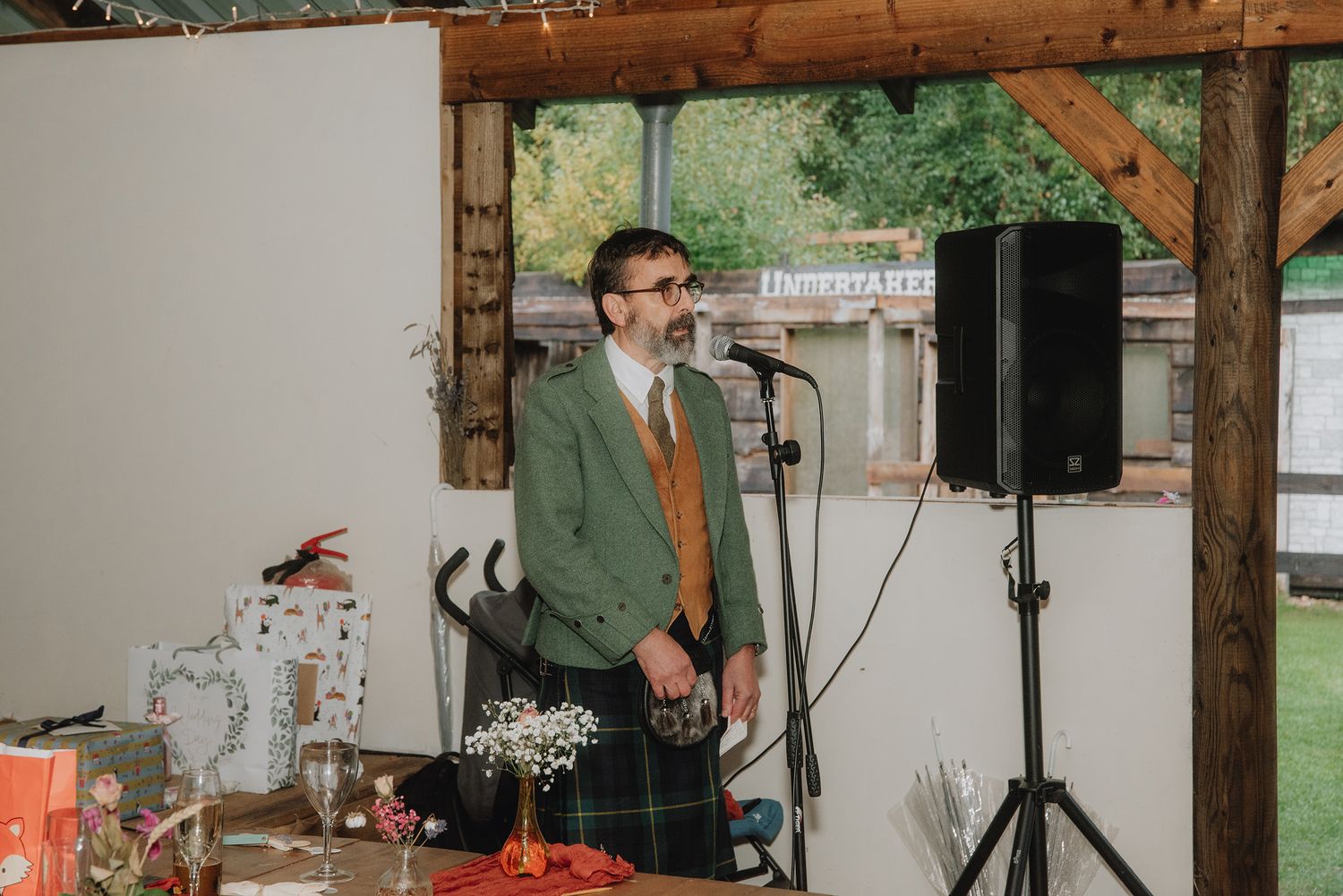 Man in green kilt and jacket giving speech at microphone during rustic wedding reception.