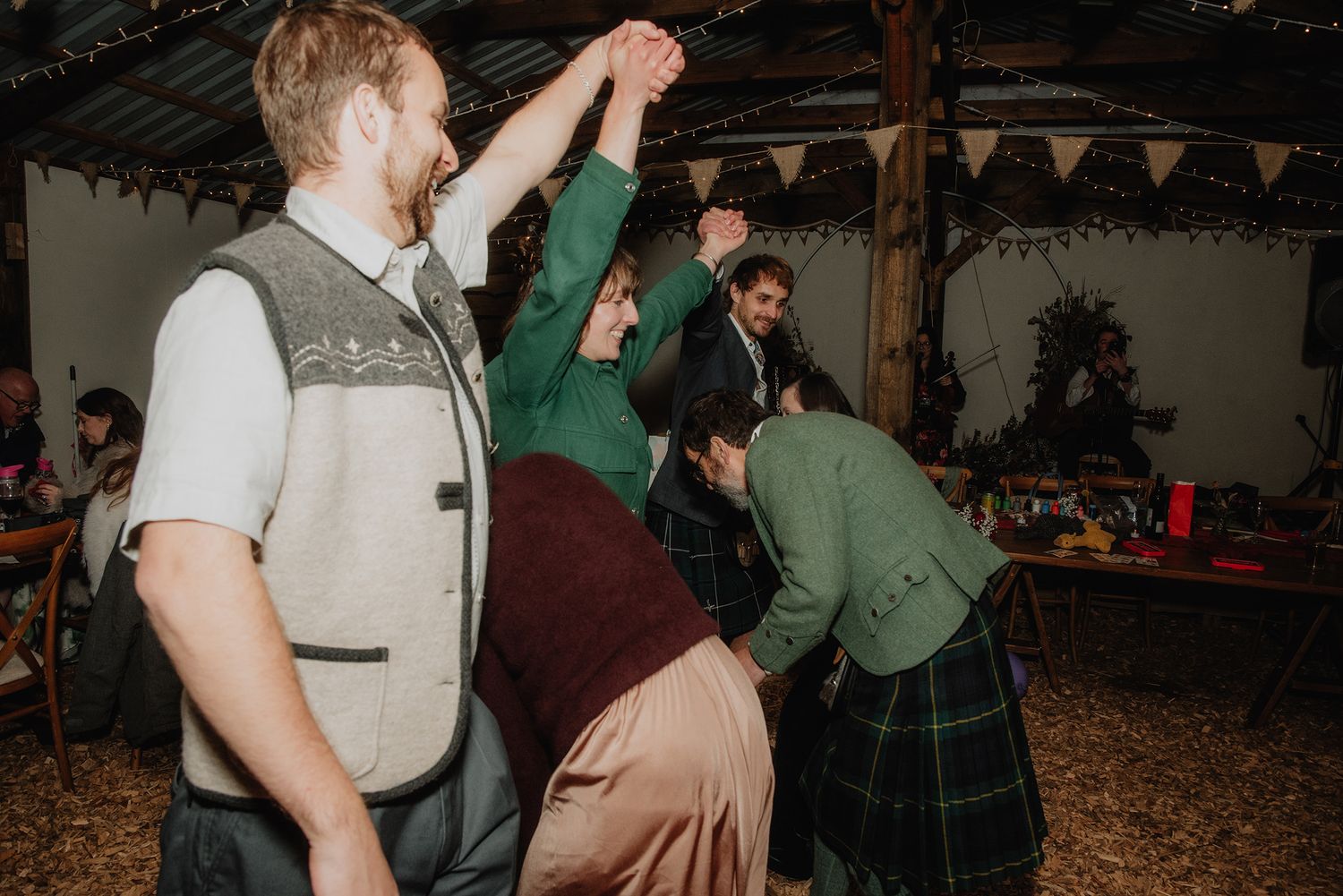 Wedding guests enjoying a lively ceilidh dance in a decorated rustic barn with string lights overhead.