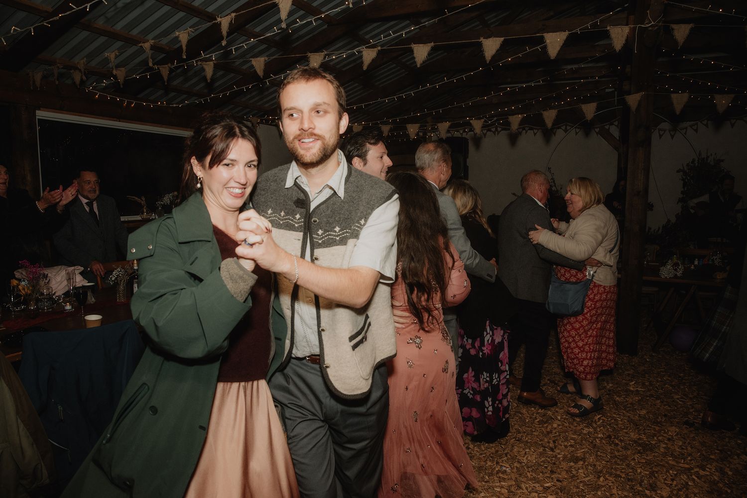 Couple smiling and dancing together at a rustic barn wedding reception with guests celebrating in the background.