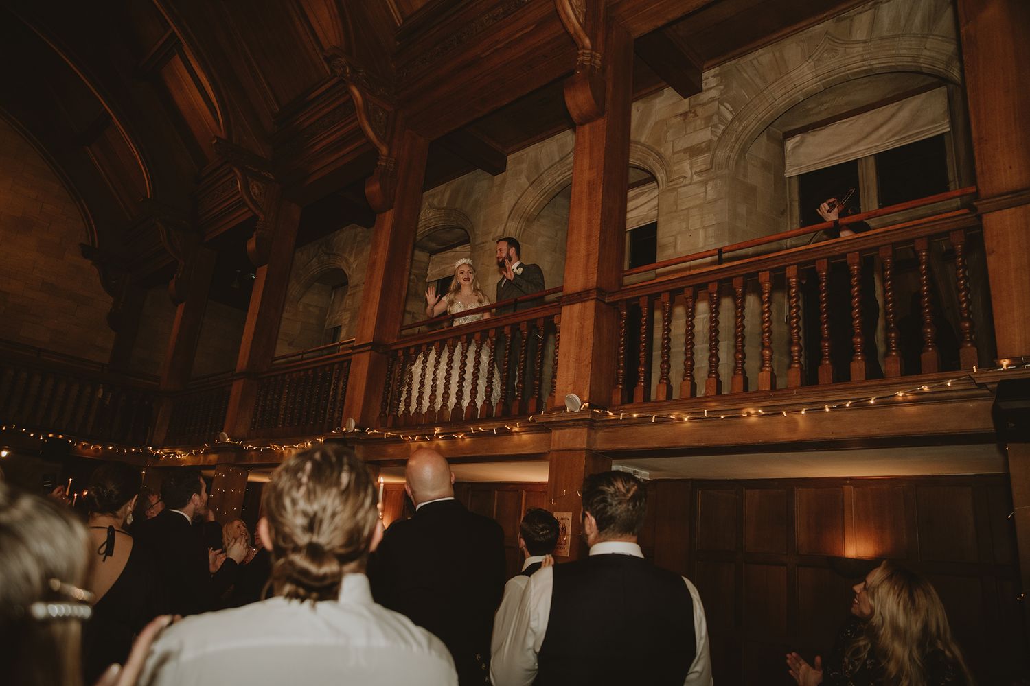 Couple speaks from a wooden balcony to wedding guests below in a rustic stone castle venue with string lights.