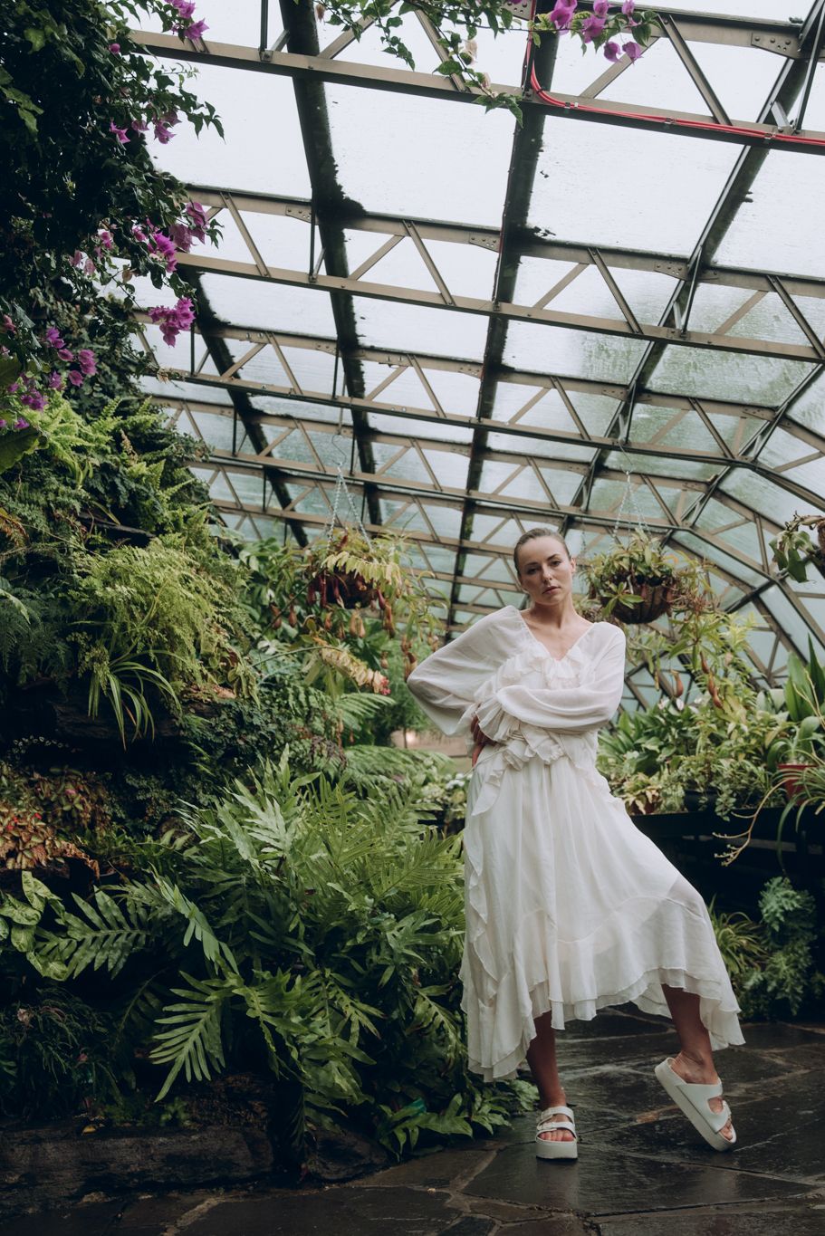 Elegant woman in flowing white dress stands in a sunlit greenhouse surrounded by tropical plants.