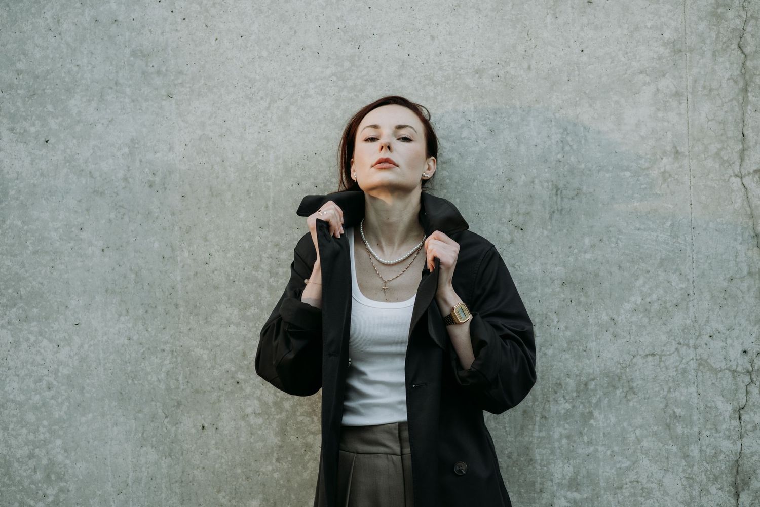 Confident woman in black jacket and white top with gold chain necklace against concrete wall.