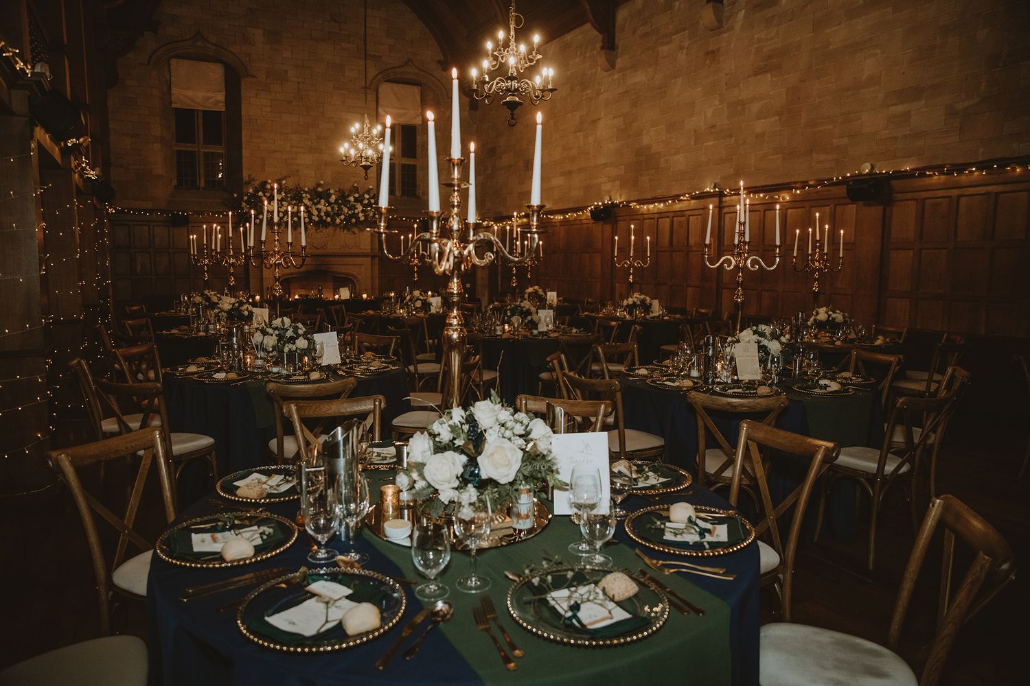 Elegant wedding reception with navy blue tablecloths, gold candelabras, white floral centerpieces, and warm candlelight in a rustic venue.