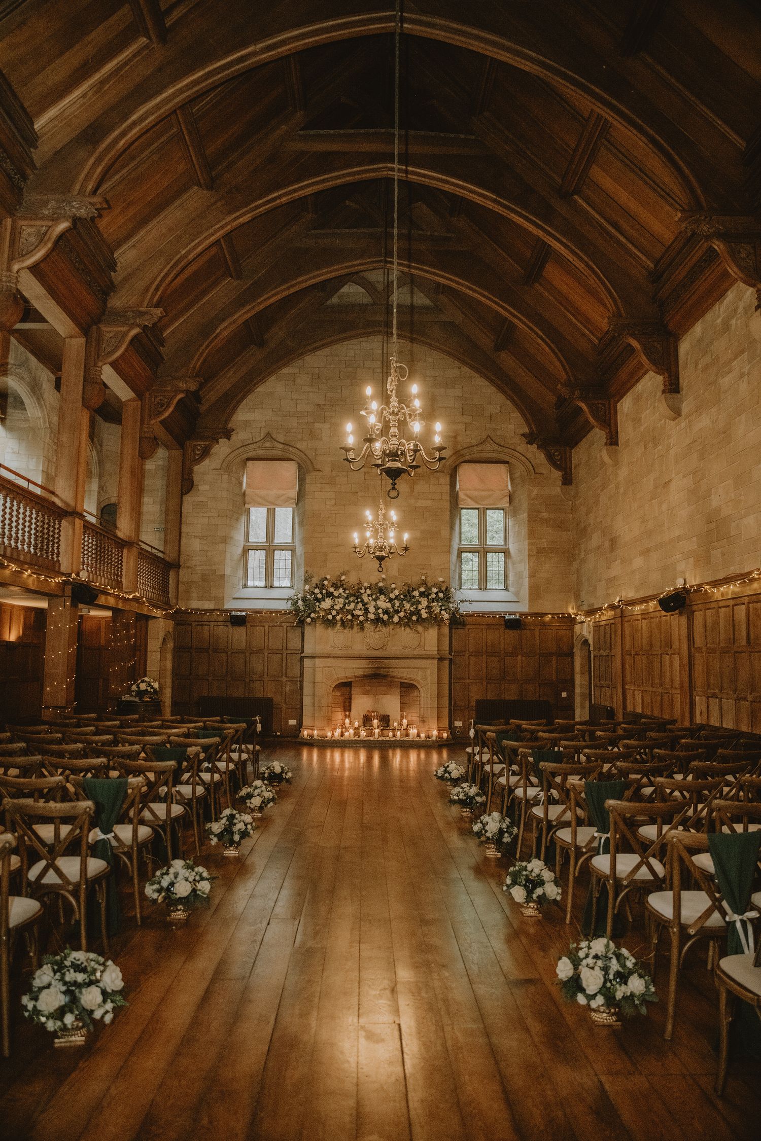 Elegant wedding ceremony hall with wooden vaulted ceiling, chandelier, floral arch over fireplace, and white flower arrangements on chairs.