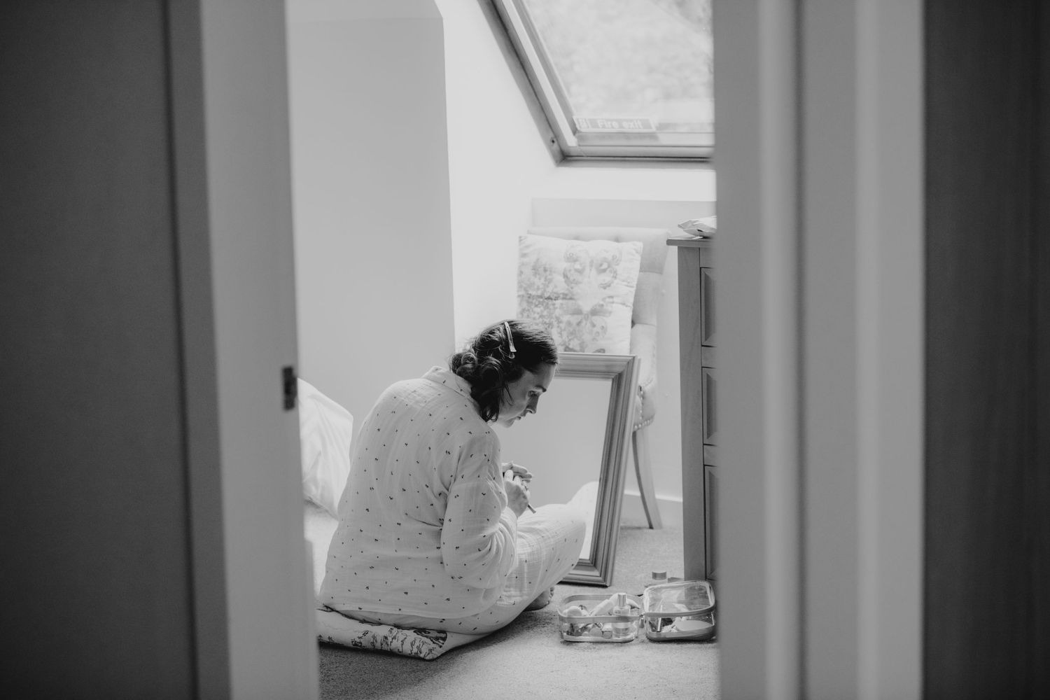 Bride in white robe sits on floor before mirror in naturally lit attic room on wedding morning.