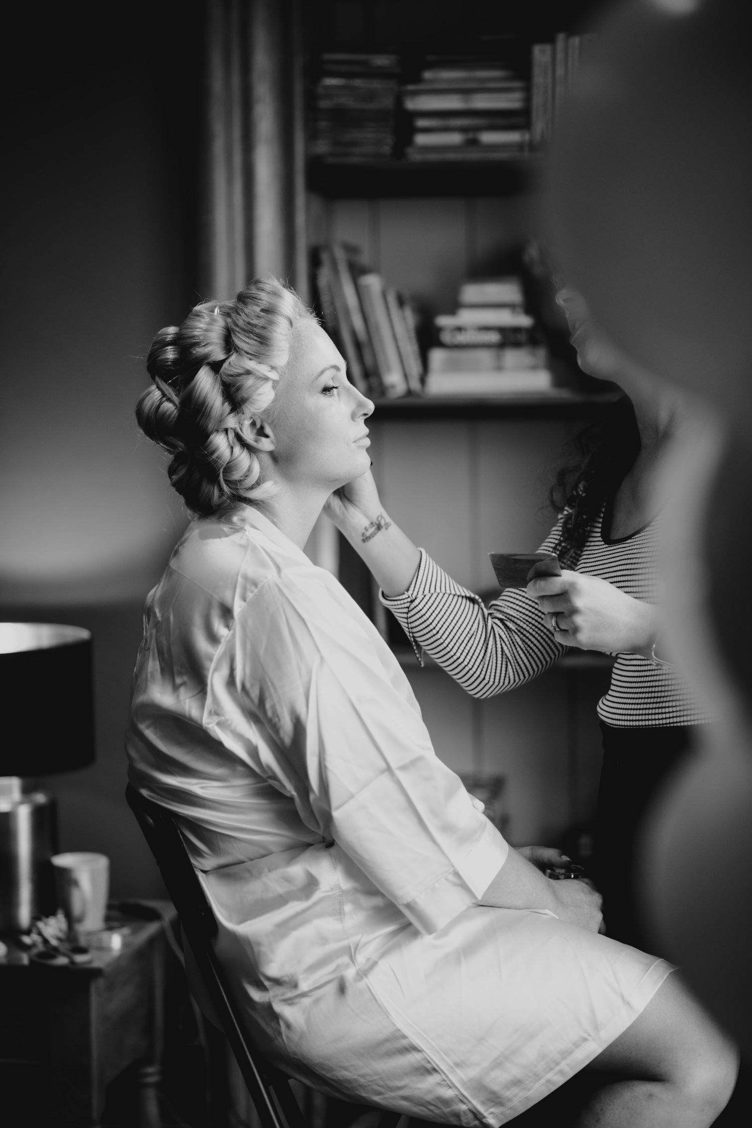 Black and white portrait of a blonde bride in a white robe having her hair styled before her wedding ceremony.