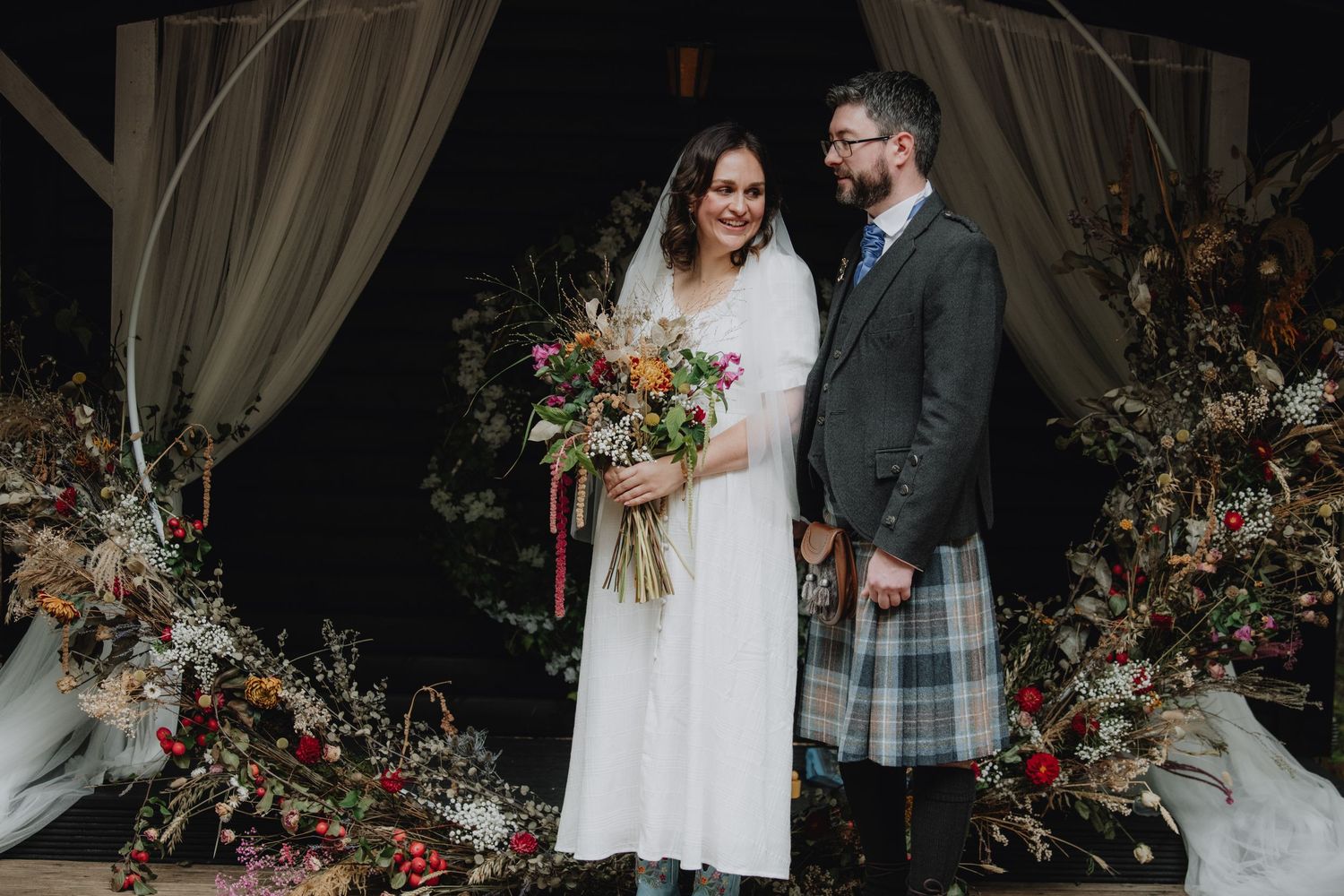 Bride and bearded groom in tartan kilt smile together at decorated woodland wedding ceremony altar.