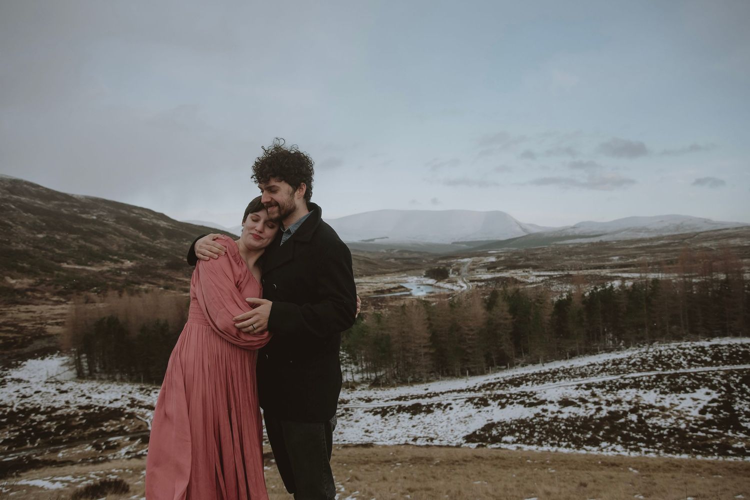 Couple embracing on a snowy Scottish highland landscape, woman in pink dress, man in black coat, misty mountains behind.