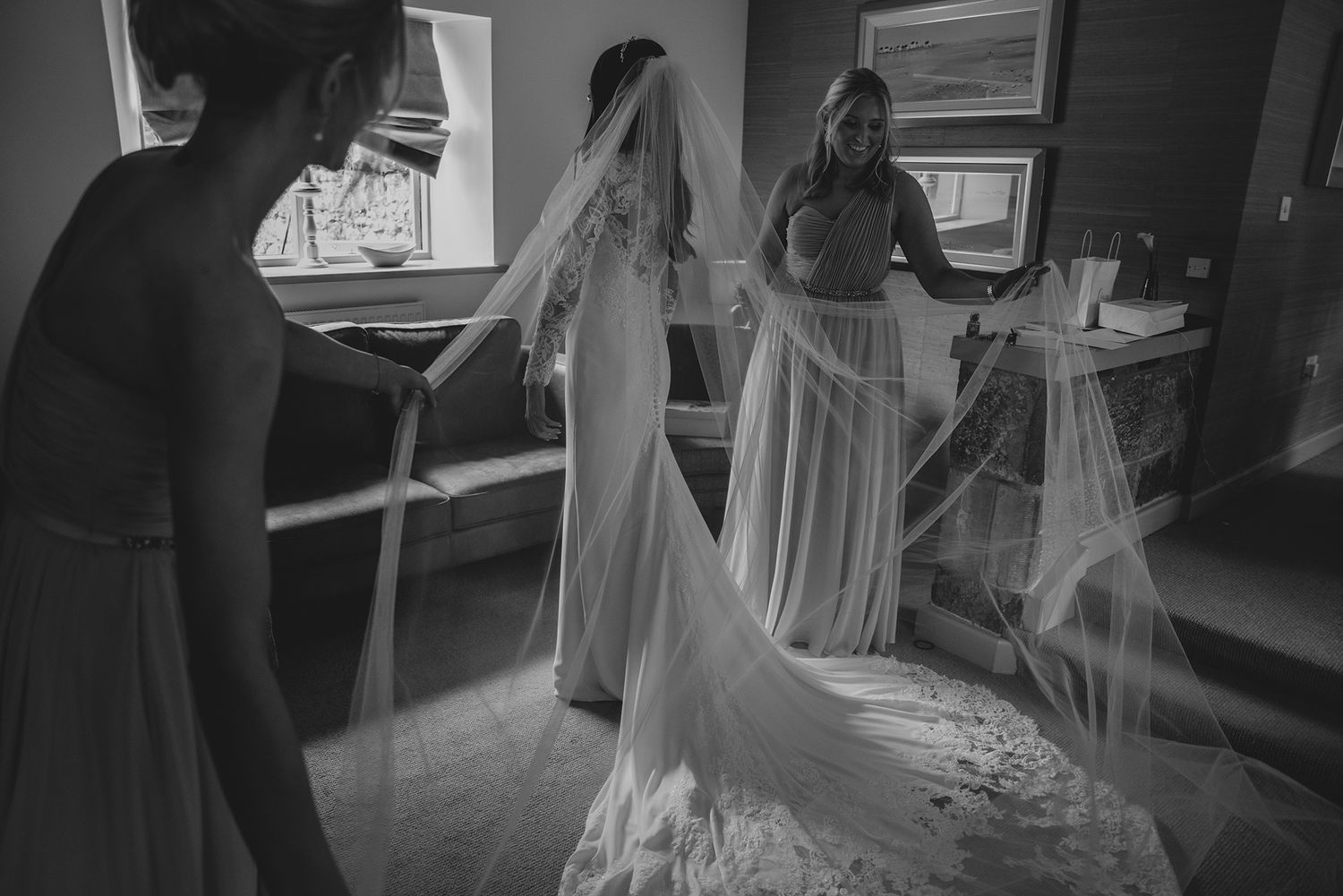 Black and white photo of a bride in a lace gown with a long veil, assisted by two bridesmaids in a bright room.