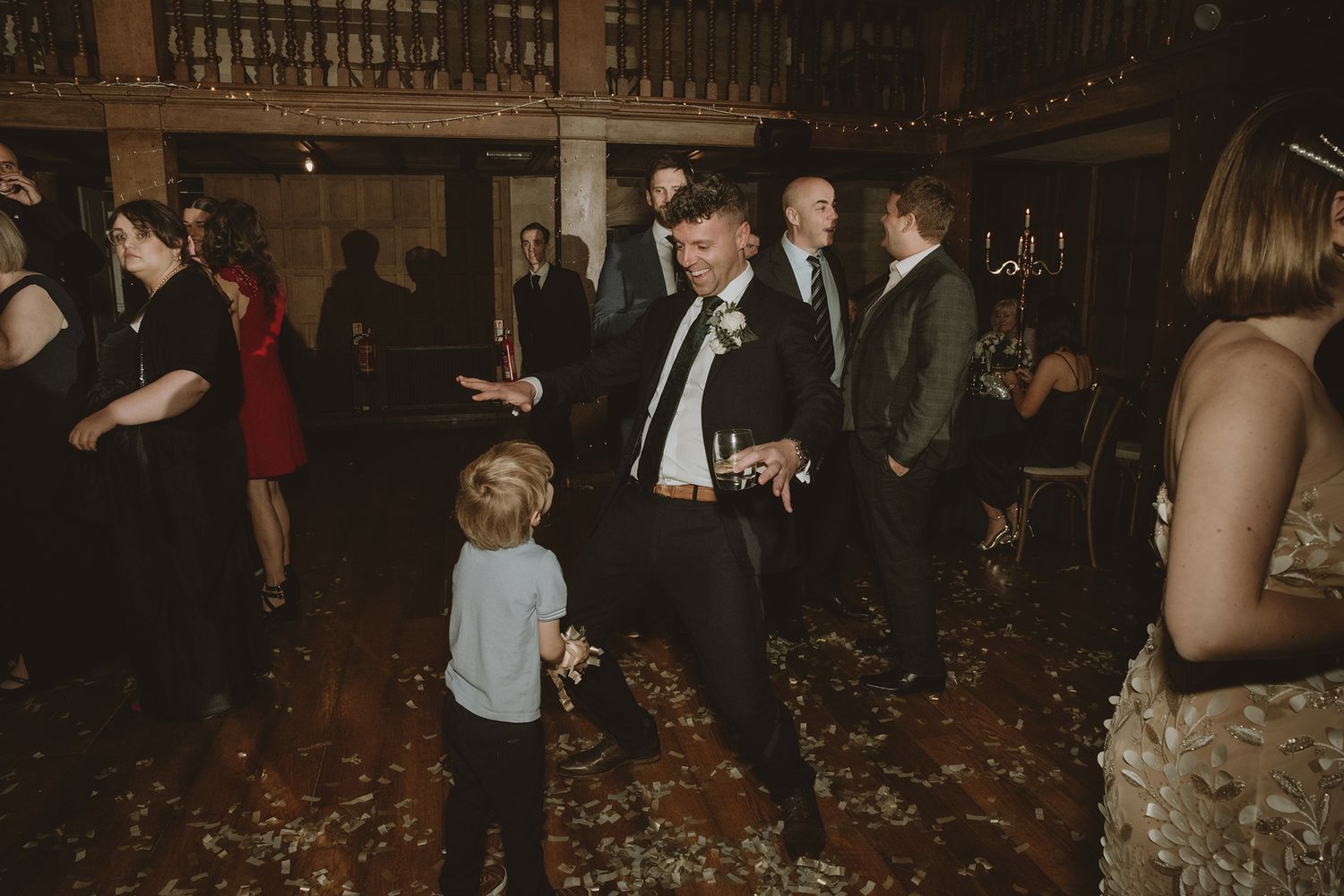 Groom in dark suit dances with young child on wooden dance floor at elegant wedding reception venue.