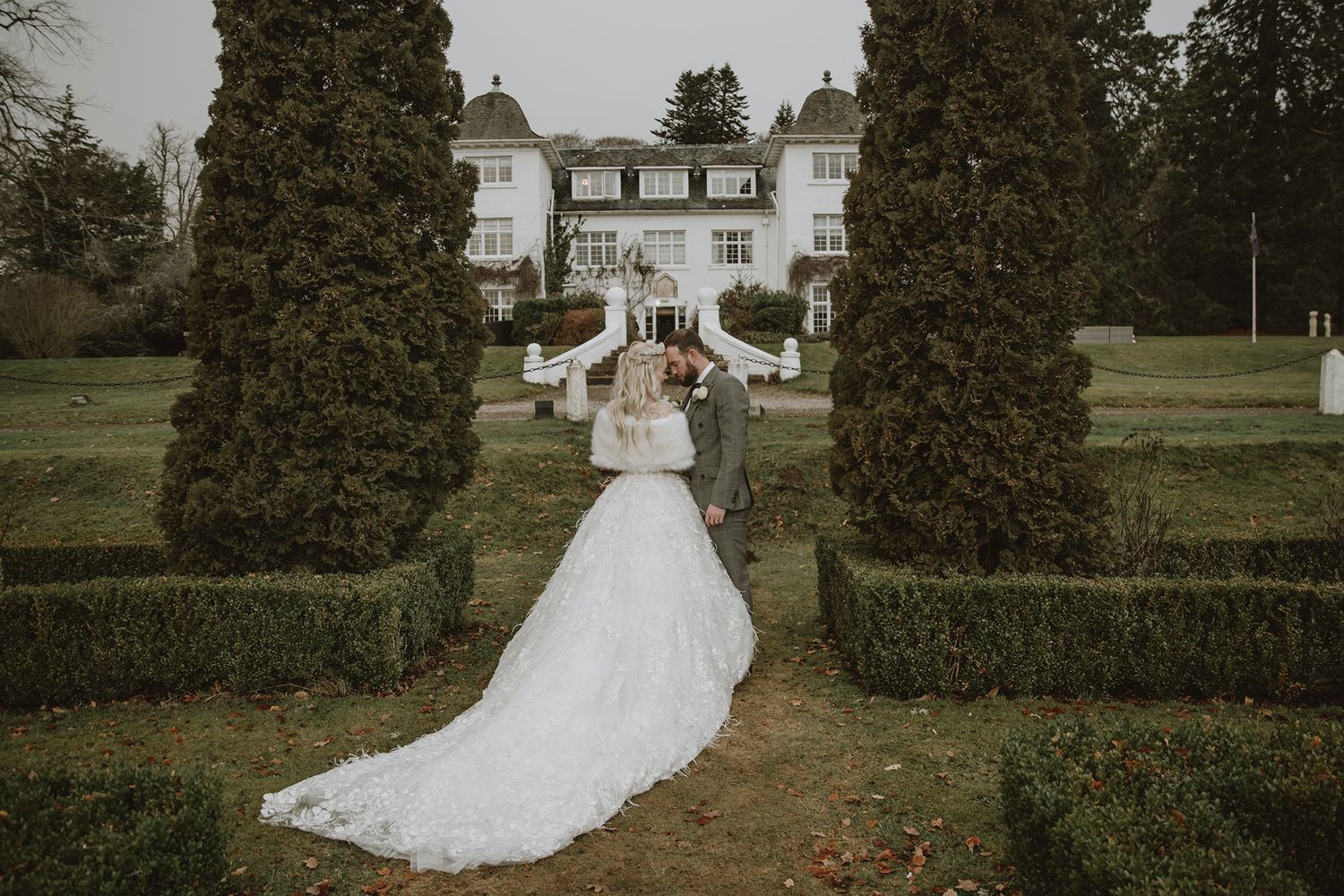 Bride and groom walk hand in hand through elegant garden with manicured hedges and grand white manor house backdrop.