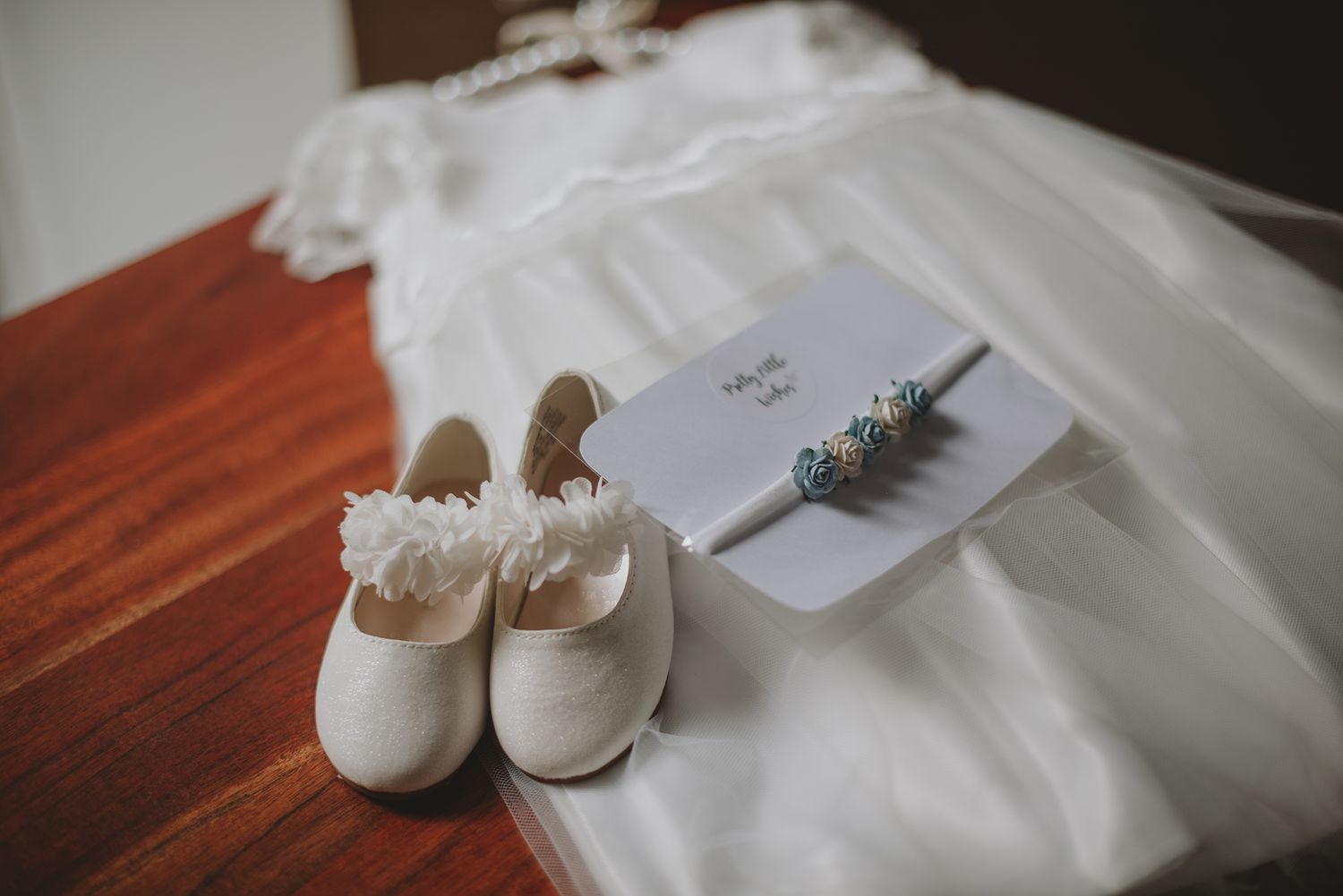 White flower girl shoes, blue floral headband, and invitation on a wooden table with white dress.