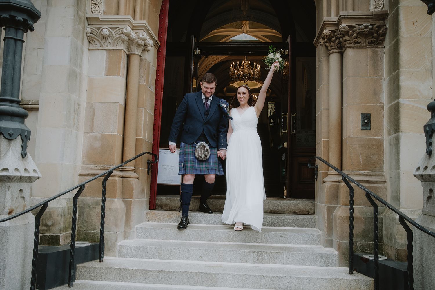 Newlyweds exit a grand stone building, bride raising bouquet in celebration, groom in traditional kilt.